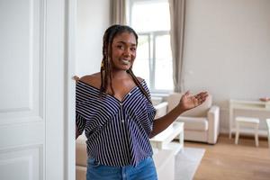 Welcome. Portrait of cheerful African woman inviting visitor to enter his home, happy young woman standing in doorway of modern apartment showing living room with hand photo