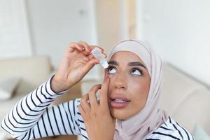 Arábica mujer con hijab utilizando ojo gota, goteante ojo lubricante a tratar seco ojo o alergia, enfermo mujer tratando globo ocular irritación o inflamación mujer sufrimiento desde irritado ojo, óptico síntomas foto