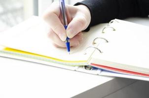 Hand holds a pen. A person writes in a notebook near the window. photo