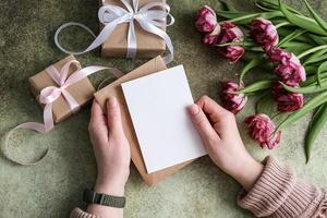 Close-up of female hands holding an envelope with an empty paper postcard on the background of a bouquet of tulips and gifts. The concept of women's day, mother's day, birthday photo