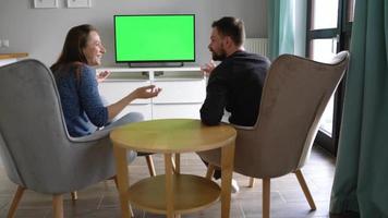 Man and woman are sitting in chairs, watching TV with a green screen, discuss what they saw and switching channels with video