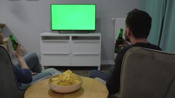Man and woman are sitting in chairs, watching TV with a green screen, drink beer and eat chips. Back view. Chroma key video