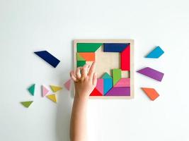 childs hand collects multicolored wooden mosaic on white background. child solves colorful tangram photo