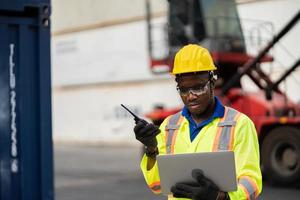 worker man in protective safety jumpsuit uniform with yellow hardhat and use laptop check container at cargo shipping warehouse. transportation import,export logistic industrial service photo