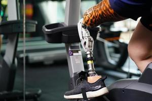 Young female with one prosthetic leg with exercising with a spinning bike in the gym to practice joint use with the other normal leg to be flexible, Concept of living a woman's life with a prosthetic photo