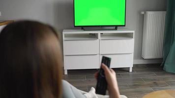 mulher é sentado dentro uma cadeira, assistindo televisão com uma verde tela, trocando canais com uma controlo remoto ao controle. croma chave video