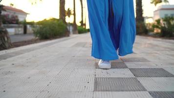 poten van een vrouw in een mooi blauw jurk rennen langs de palm Laan. hoed valt naar de grond. dramatisch langzaam beweging video