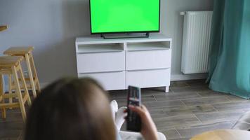 mujer es sentado en un silla, acecho televisión con un verde pantalla, traspuesta canales con un remoto control. croma llave video