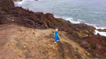 ver desde el altura de mujer en un hermosa azul vestir y sombrero soportes en parte superior de un montaña en un conservación zona en el costas de el atlántico océano. tenerife, canario islas, España video