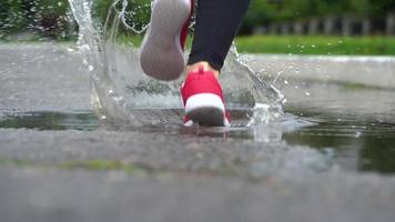 stänga upp av ben av en löpare i sneakers. sporter kvinna joggning utomhus, stepping in i grumlig pöl. enda löpare löpning i regn, framställning stänk video