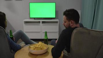 Man and woman are sitting in chairs, watching TV with a green screen, drink beer and eat chips. Back view. Chroma key video