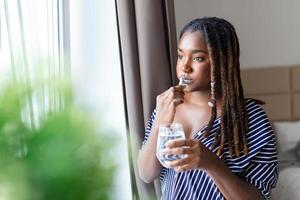 Young African woman brushing teeth with toothbrush, holding glass of water and looking in window photo