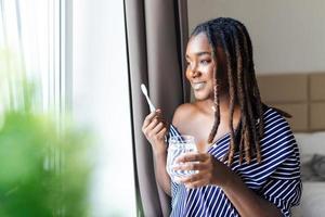 joven africano mujer cepillado dientes con cepillo de dientes, participación vaso de agua y mirando en ventana foto