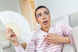 Stressed annoyed woman using waving fan suffer from overheating, summer heat health hormone problem, no air conditioner at home sit on sofa feel exhaustion dehydration heatstroke concept photo