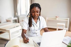 African professional female doctor wearing uniform taking notes in medical journal, filling documents, patient illness history, looking at laptop screen, student watching webinar photo