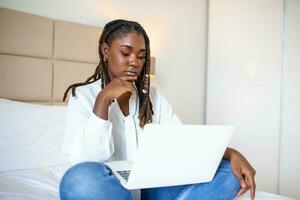 Attractive African woman in white shirt using laptop and smiling at morning. Video chat and freelance working from home concept of beautiful young woman with laptop in bed photo