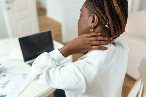 retrato de joven estresado africano mujer sentado a hogar oficina escritorio en frente de computadora portátil, conmovedor dolor hombro con dolorido expresión, sufrimiento desde hombro dolor después trabajando en ordenador personal foto