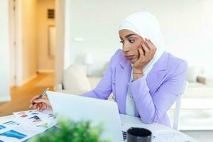 trastornado mujer vistiendo hijab en frente de ordenador portátil buscar y haciendo oficina trabajar, negocio, Finanzas y puesto de trabajo concepto. árabe mujer de negocios trabajando a hogar. foto