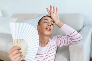 cansado mujer sufrimiento desde golpe de calor o caliente verano plano sin aire acondicionado, utilizando ondulación admirador, sentado en vivo habitación a hogar. calentamiento excesivo foto