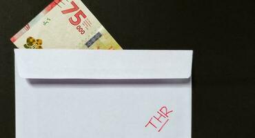 A white envelope written of THR and new Indonesian banknotes, usually Tunjangan Hari Raya or called THR are given to employees ahead of Eid. Isolated on black background and top view photo