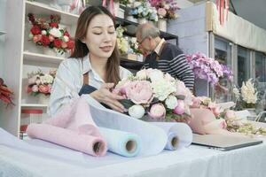 Young beautiful Asian female florist entrepreneur arranging a bunch of blossoms works with old man shopkeeper, happy work in colorful flower shop store with fresh blooms, small business, family SME. photo