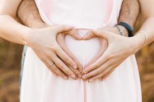 el futuro padre abrazos el barriga de un embarazada mujer, el expectante madre y hace manos con su en el formar de un corazón. cuidado y amor para el niño foto