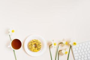 Workplace of a flat lay blogger or freelancer is a woman with a keyboard, a cup of tea, donuts on a plate and spring flowers daffodils on a light background with a copy space photo