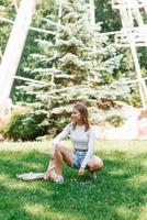 Young Caucasian woman in denim shorts and a white top sat down on the grass in an amusement park photo
