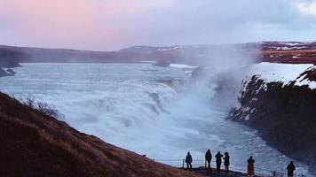 Tourist visit stand on viewpoint Gullfoss waterfall in Iceland, Cinematic beautiful majestic winter waterfall cover by snow and ice. Slow motion. Purple Sunset sky video