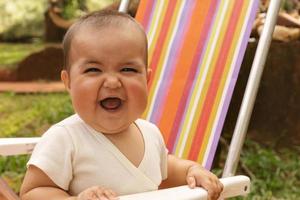 de cerca de bebé niña riendo y mirando a el cámara, sentado en un vistoso silla al aire libre. foto