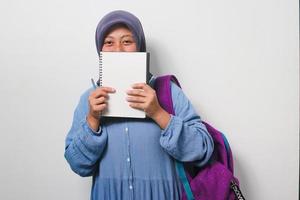 Young Asian girl student in hijab covering her face with book isolated on white background. photo