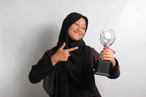 inteligente joven asiático niña estudiante en hijab señalando su trofeo aislado en blanco antecedentes. foto