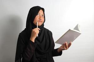 Thoughtful young Asian girl student interested in thinking while holding a book and pen isolated on white background photo