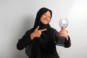 Clever young Asian girl student in hijab pointing her trophy isolated on white background. photo