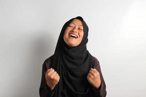 Excited young Asian girl in hijab raising fist isolated on white background. photo