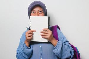 Young Asian girl student in hijab covering her face with book isolated on white background. photo