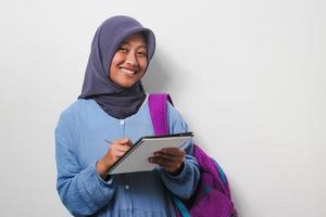 inteligente joven asiático niña estudiante en hijab participación un bolígrafo y libro en blanco antecedentes. foto