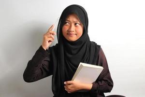 Thoughtful young Asian girl student interested in thinking while holding a book and pen isolated on white background photo