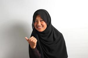 Cheerful young Asian girl in hijab pointing to the side with thumbs up isolated on white background. photo