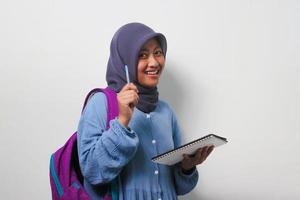 inteligente joven asiático niña estudiante en hijab participación un bolígrafo y libro en blanco antecedentes. foto