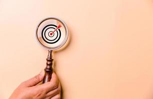Hand holding target board inside vintage magnifying glass for focus business objective on orange background and copy space. Search, goal, strategy, success. successful marketing strategy goals photo