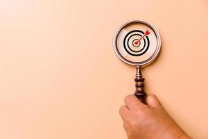 Hand holding target board inside vintage magnifying glass for focus business objective on orange background and copy space. Search, goal, strategy, success. successful marketing strategy goals photo
