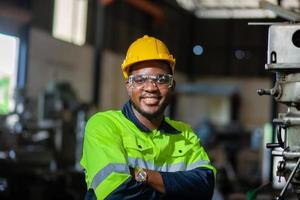 retrato de un africano americano ingeniero en pie con su brazos cruzado, inspeccionando y controlador un automatizado soldadura robot. el robótico brazo en el automotor fábrica es elegante. foto