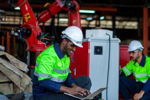 africano americano fábrica trabajador trabajando con un robótico brazo en un fábrica. industrial robot antecedentes para automatizado producción tecnología. mirando a cámara foto