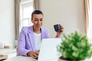 Remote job, technology and people concept - happy smiling young black business woman with laptop computer and papers working at home office photo