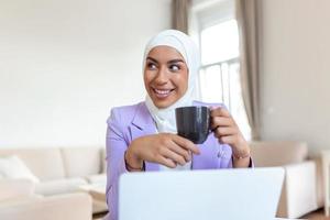 Muslim woman working with computer. Arab Young business woman sitting at her desk at home, working on a laptop computer and drinking coffee or tea. Muslim woman working at a home and using computer. photo