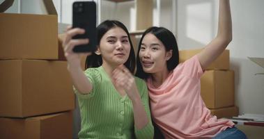 métrage de content asiatique Jeune femmes vidéo appel salutation avec téléphone intelligent tandis que séance sur le sol dans Nouveau appartement. en mouvement et mode de vie concepts. video