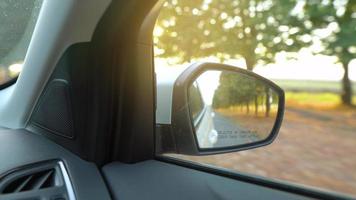 Visão a partir de a visão traseira espelho Como carro drives através a estrada cercado de árvores às pôr do sol video