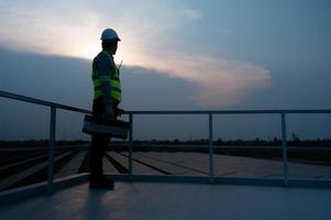 Electrical engineers inspecting solar panels in a hundred acres of grass on the rooftop of energy storage station, in the evening after completing the daily work tasks with the setting sun. photo