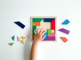 childs hand collects multicolored wooden mosaic on white background. child solves colorful tangram photo